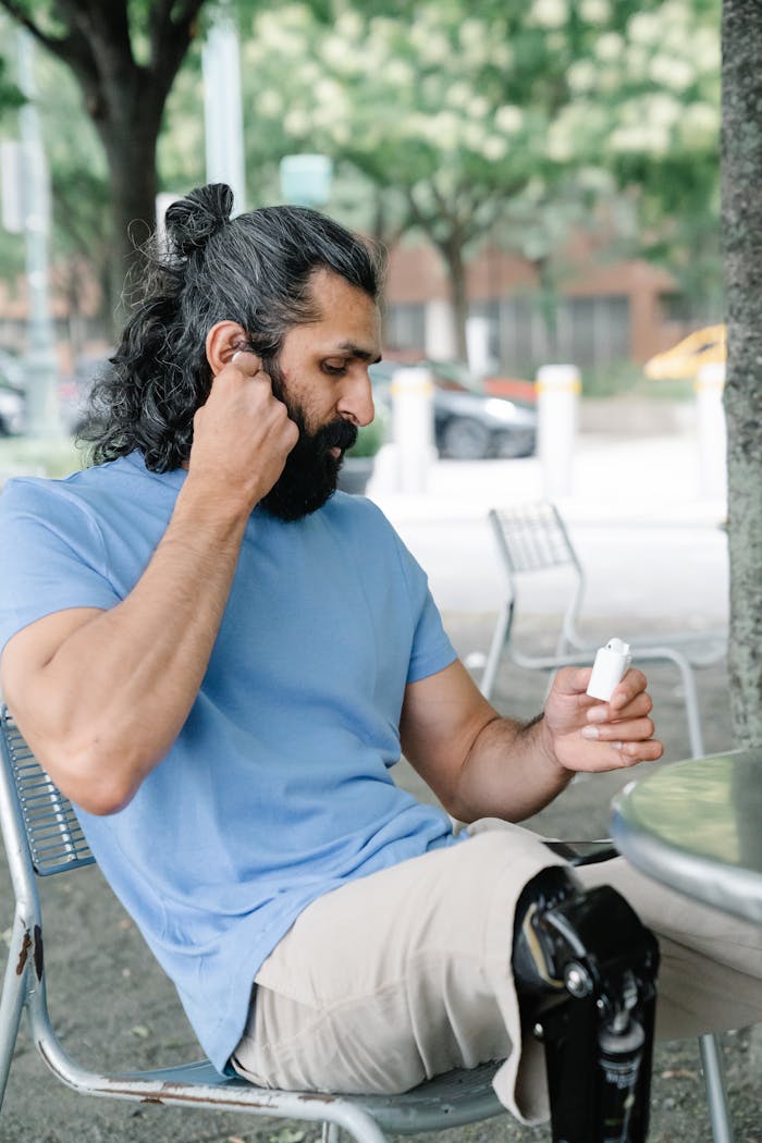 about-04 Man adjusting hearing aid while seated at a park table, showcasing body positivity and disability awareness.